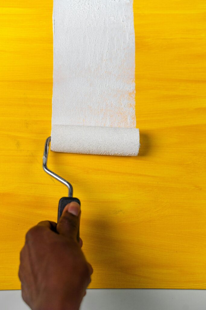 A hand using a roller to apply white paint on a yellow canvas.